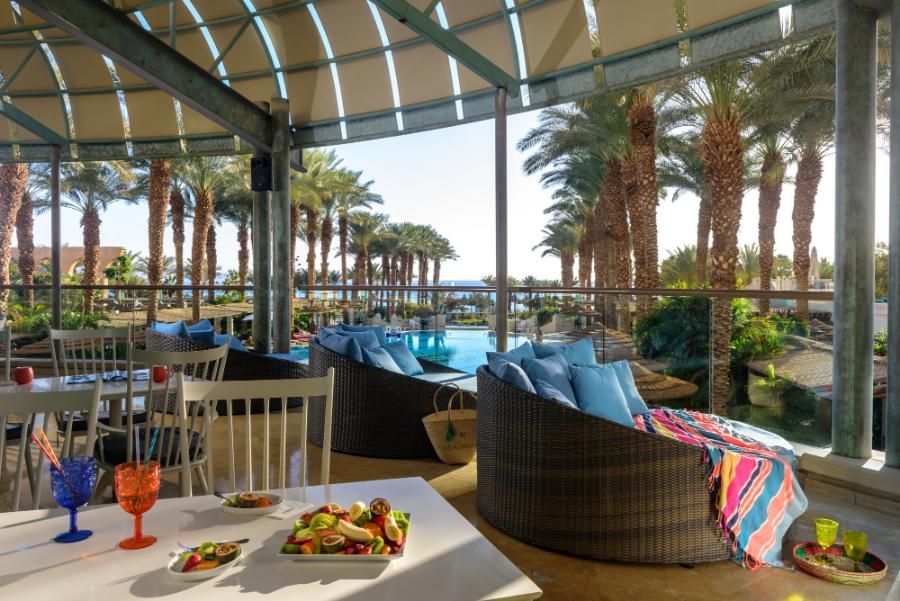פינת ישיבה בטרסה במלון הרודס פאלאס אילת, עם נוף לבריכה ודקלים, שולחנות ערוכים עם פירות וקוקטיילים