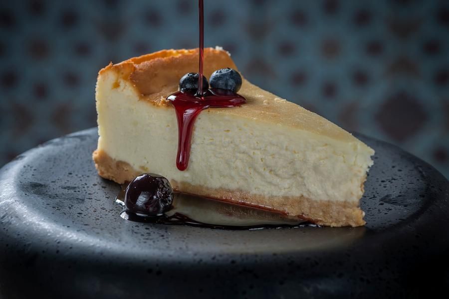 פרוסת עוגת גבינה אפויה עם אוכמניות, מוגשת עם זיגוג רוטב פירות יער, על צלחת כהה