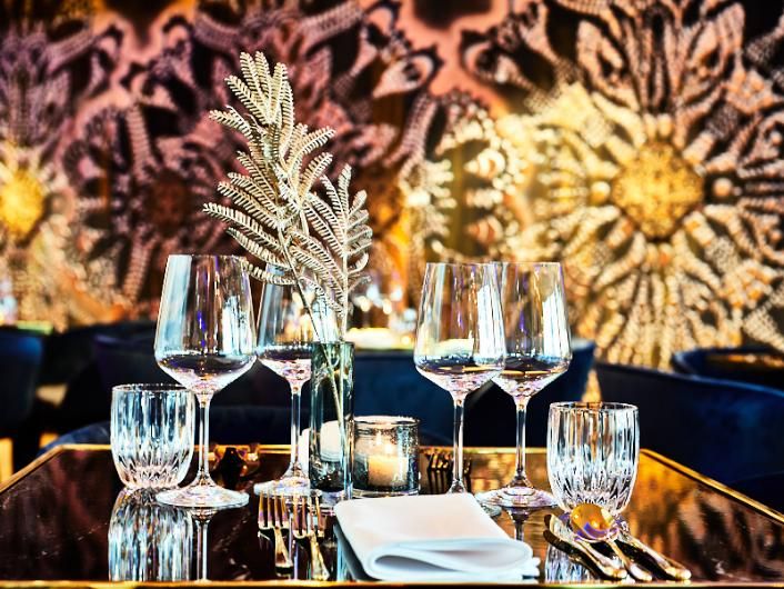 שולחן זכוכית שחור עם כוסות יין, כלי זכוכית, סכו"ם ומפית לבנה, על רקע קיר שחור עם דקורציה מוזהבת 