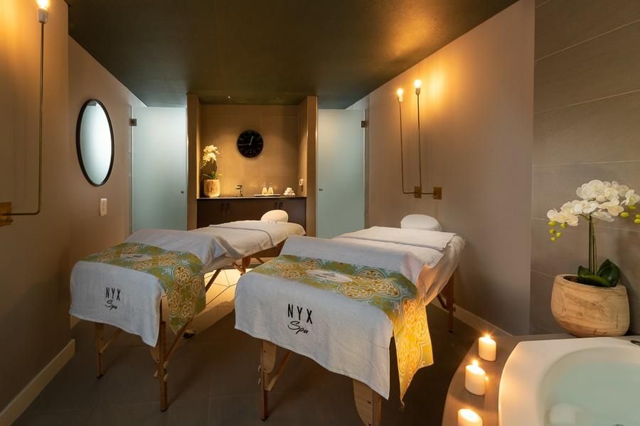 חדר טיפולים זוגי בספא NYX, עם שני מיטות עיסוי מכוסות במגבות לבנות ובד צבעוני ונרות דולקים ליד ג'קוזי