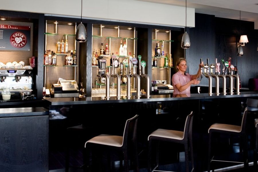 בר המלון לאונרדו דבלין קרייסטצ'רץ' עם דלפק שחור, ברזי בירה יוקרתיים וברמנית מוזגת משקה