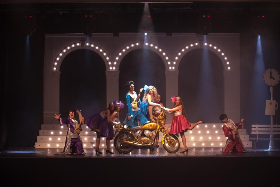 מופע תיאטרון מוזיקלי עם שחקנים בתלבושות צבעוניות ואופנוע זהוב על במה מוארת